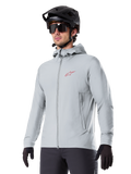 Veste thermique Alpinestars A-Dura, veste de cyclisme isolante gris clair, design à capuche avec petit logo rouge, portée avec un casque et des gants noirs, vêtement technique de VTT pour rouler par temps froid