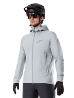 Veste thermique Alpinestars A-Dura, veste de cyclisme isolante gris clair, design à capuche avec petit logo rouge, portée avec un casque et des gants noirs, vêtement technique de VTT pour rouler par temps froid
