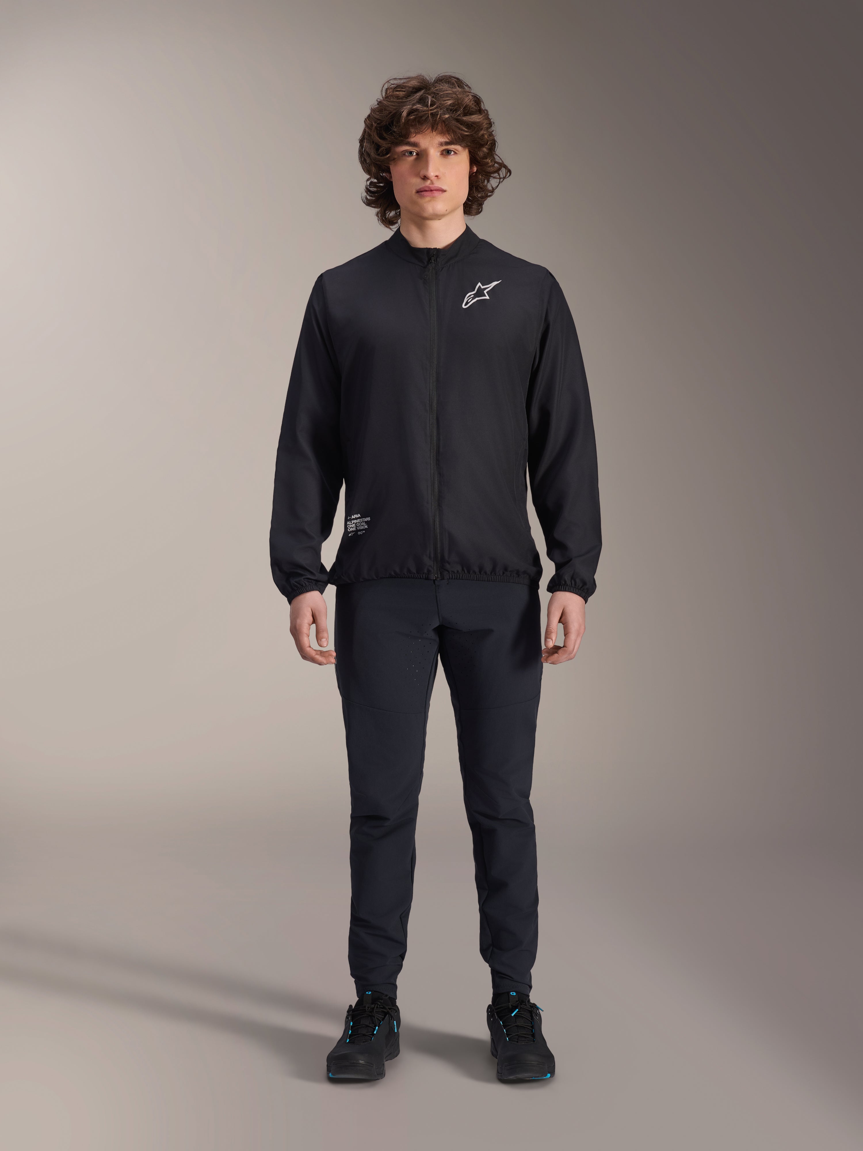 Modèle portant la veste coupe-vent Alpinestars A-Aria en noir, dotée d'une fermeture éclair intégrale, d'un col montant et d'un logo Astars blanc sur la poitrine, associée à un pantalon technique noir et des chaussures noires, vêtement de cyclisme performant pour le VTT et l'extérieur