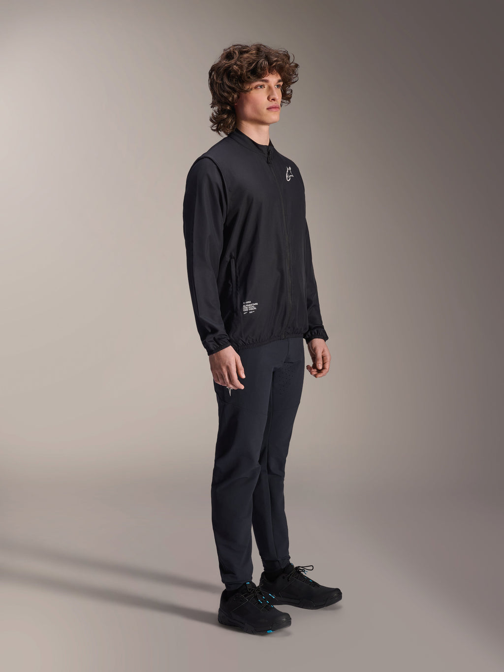 Veste coupe-vent Alpinestars A-Aria, noire, veste de cyclisme légère et résistante au vent portée par un mannequin aux cheveux bouclés, avec un logo Astars blanc sur la poitrine et un marquage en bas à droite, associée à un pantalon et des chaussures noirs, coupe performance pour le VTT et le gravel