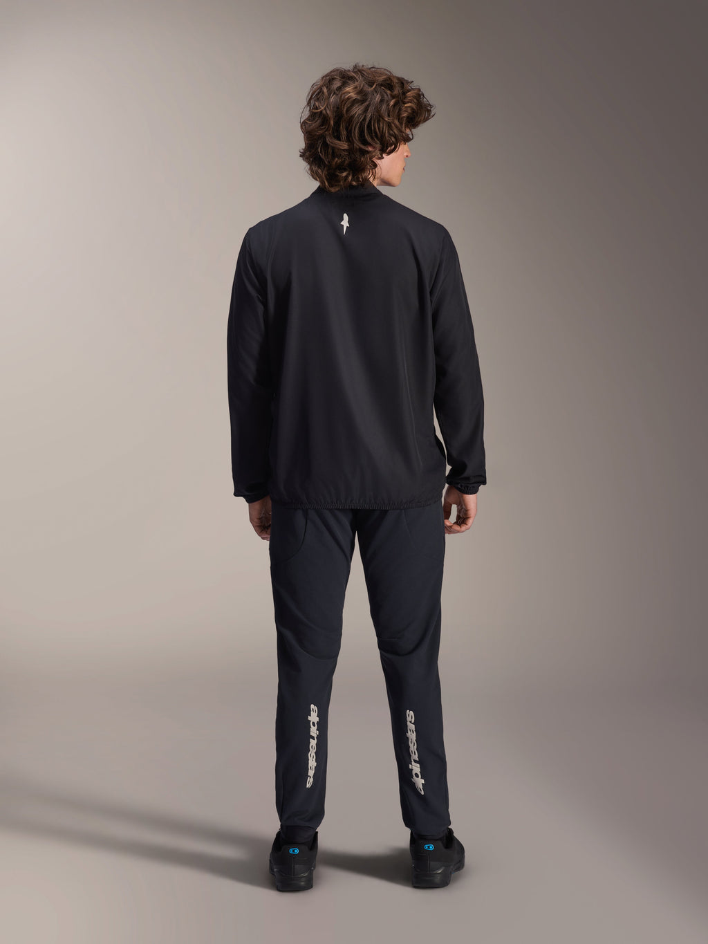 Veste coupe-vent Alpinestars A-Aria, noire, vue de dos, veste de cyclisme légère et résistante au vent avec un petit logo blanc sur le haut du dos, portée avec un pantalon noir assorti arborant le marquage vertical Alpinestars sur les mollets, équipement de VTT haute performance