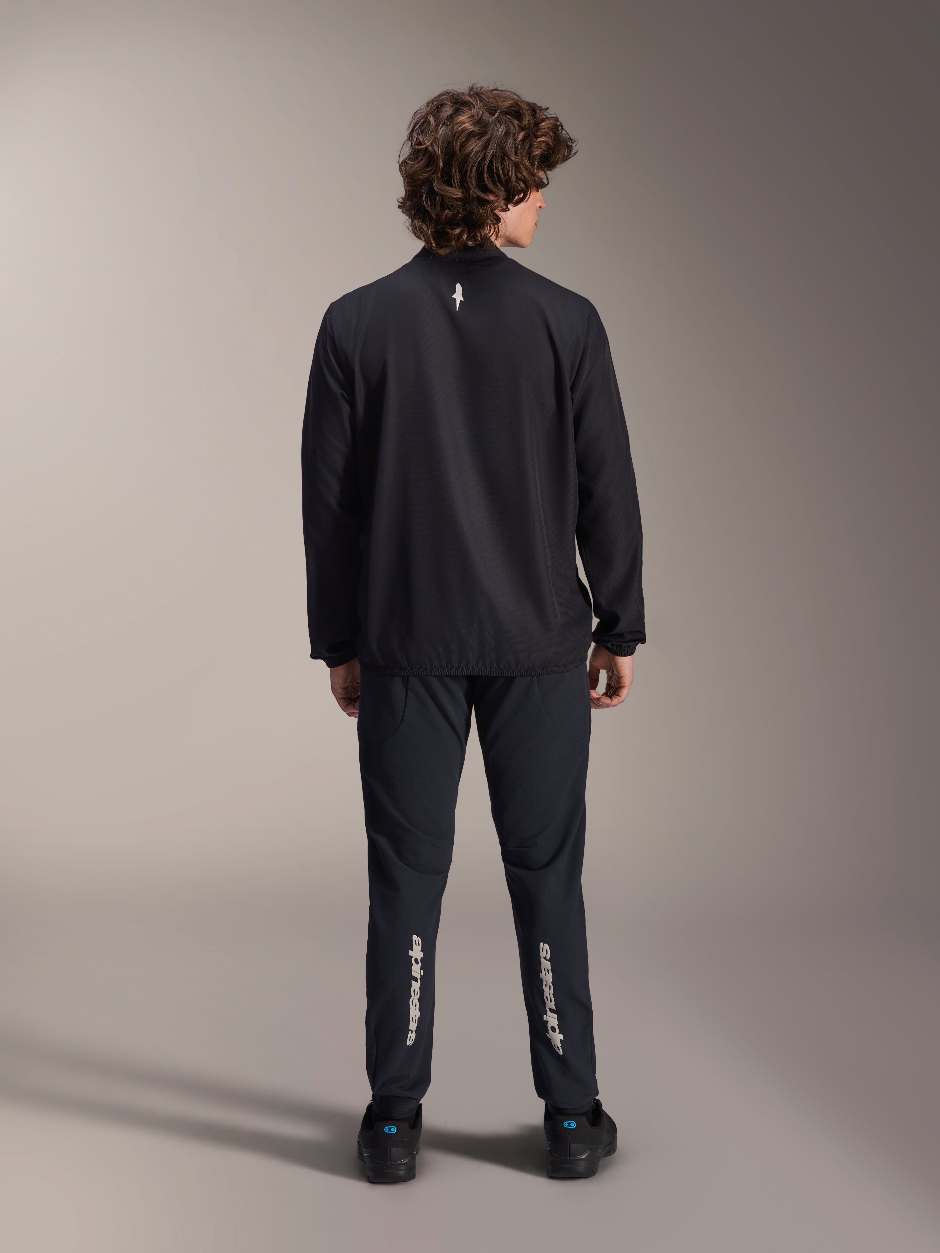 Veste coupe-vent Alpinestars A-Aria, noire, vue de dos, veste de cyclisme légère et résistante au vent avec un petit logo blanc sur le haut du dos, portée avec un pantalon noir assorti arborant le marquage vertical Alpinestars sur les mollets, équipement de VTT haute performance