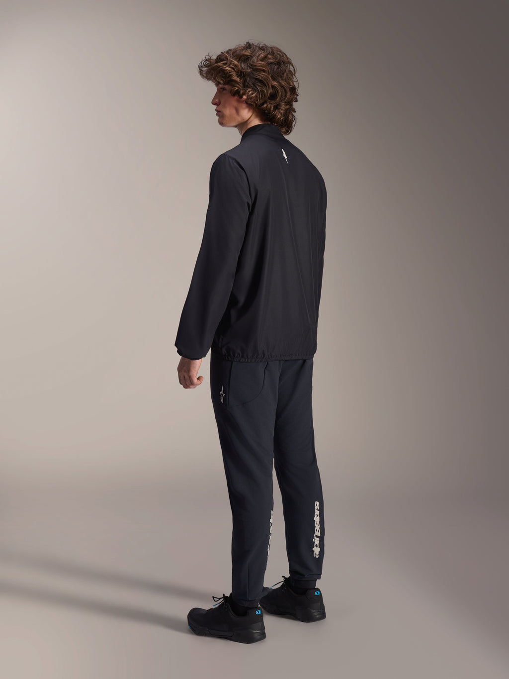 Veste coupe-vent Alpinestars A-Aria, noire, veste de cyclisme légère et résistante au vent, vue de trois-quarts arrière sur modèle, dotée d'un petit logo blanc au niveau de la nuque et d'un ourlet élastiqué, associée à un pantalon et des chaussures noirs assortis pour un look axé sur la performance.