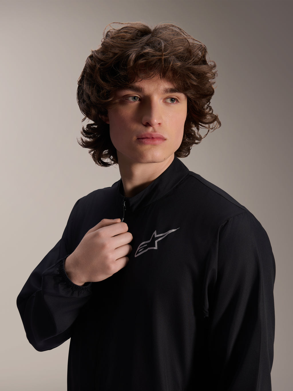 Veste coupe-vent Alpinestars A-Aria, veste de cyclisme noire, portée par un mannequin homme, col montant avec logo Astars argenté sur la poitrine, équipement de VTT performant, léger et résistant au vent
