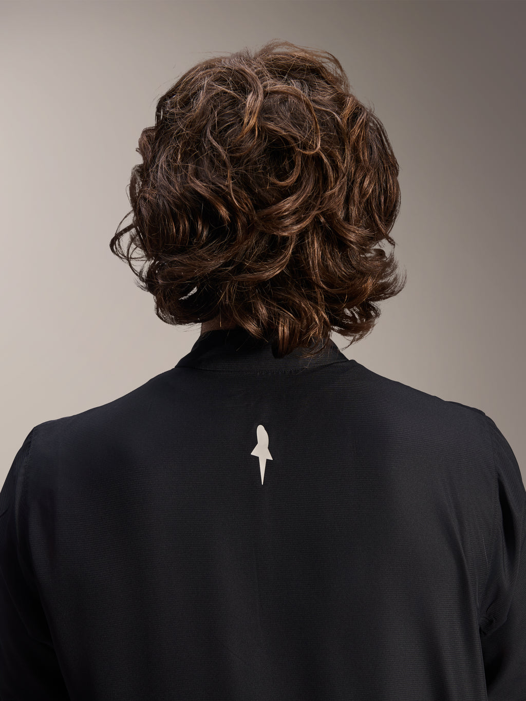 Veste coupe-vent Alpinestars A-Aria, noire, vue rapprochée de dos du haut des épaules et du col, avec un petit logo blanc au centre du dos, portée par un mannequin aux cheveux bruns ondulés, équipement de cyclisme technique et minimaliste