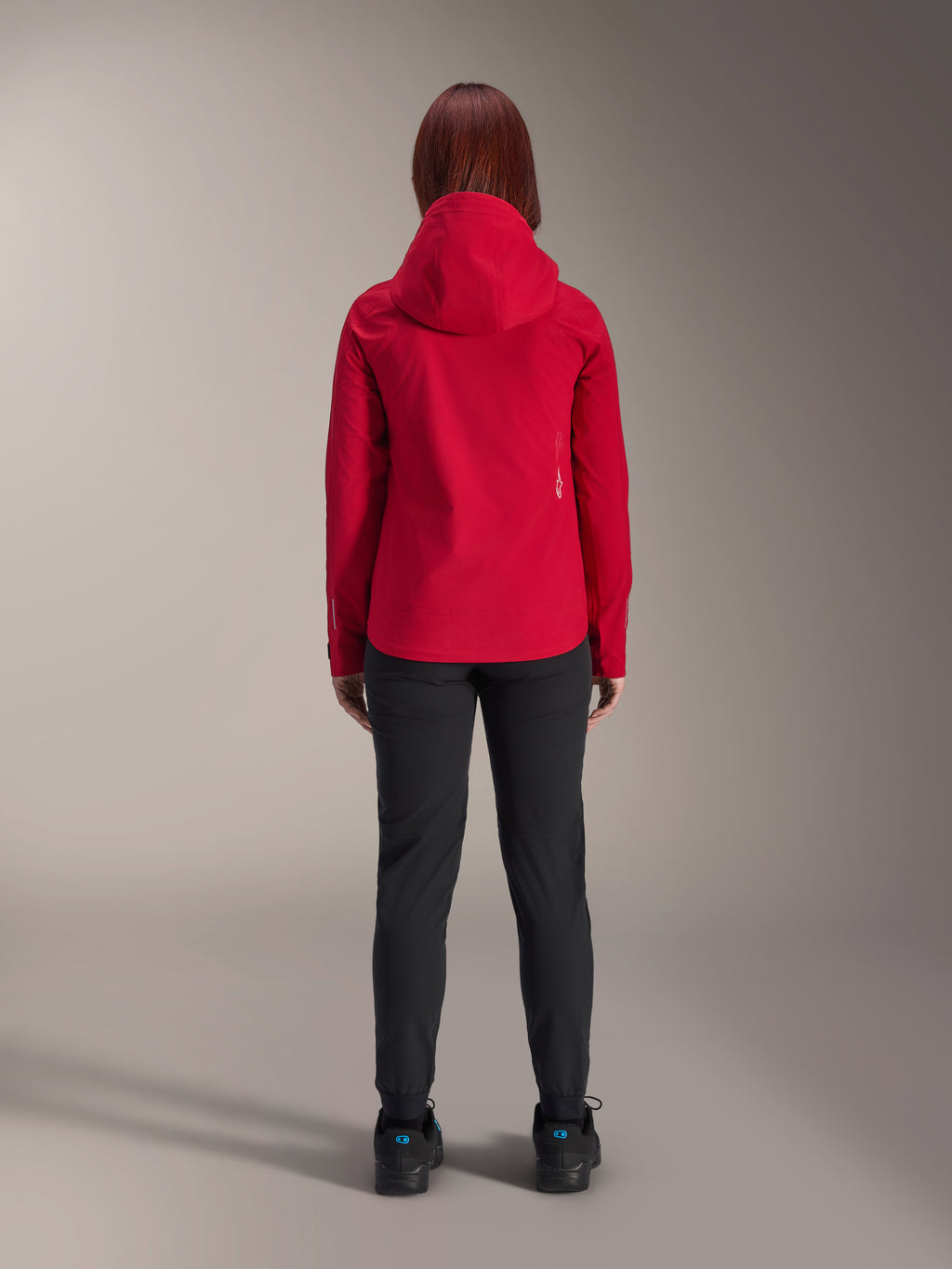 Back view of a woman wearing the Alpinestars Stella A-Dura Elite Waterproof Jacket in Red Fluo, styled with black pants and shoes, featuring a protective hood and dropped tail hem for mountain biking.