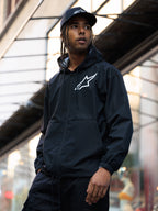 Veste coupe-vent Alpinestars Speeder Chromium, noire, coupe-vent à capuche avec logo blanc sur la poitrine, porté par un homme avec une casquette Alpinestars noire assortie dans un environnement urbain, style streetwear motorsport décontracté
