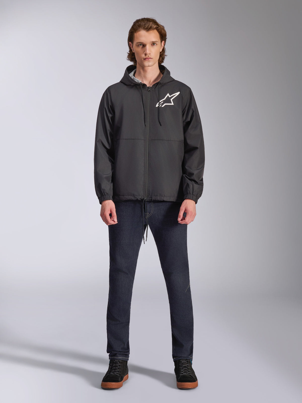 Veste coupe-vent Alpinestars Speeder Chromium, noire avec logo Astars blanc, portée par un mannequin avec un jean bleu foncé et des baskets noires, dotée d'une capuche et d'une fermeture éclair frontale, streetwear décontracté inspiré du sport automobile