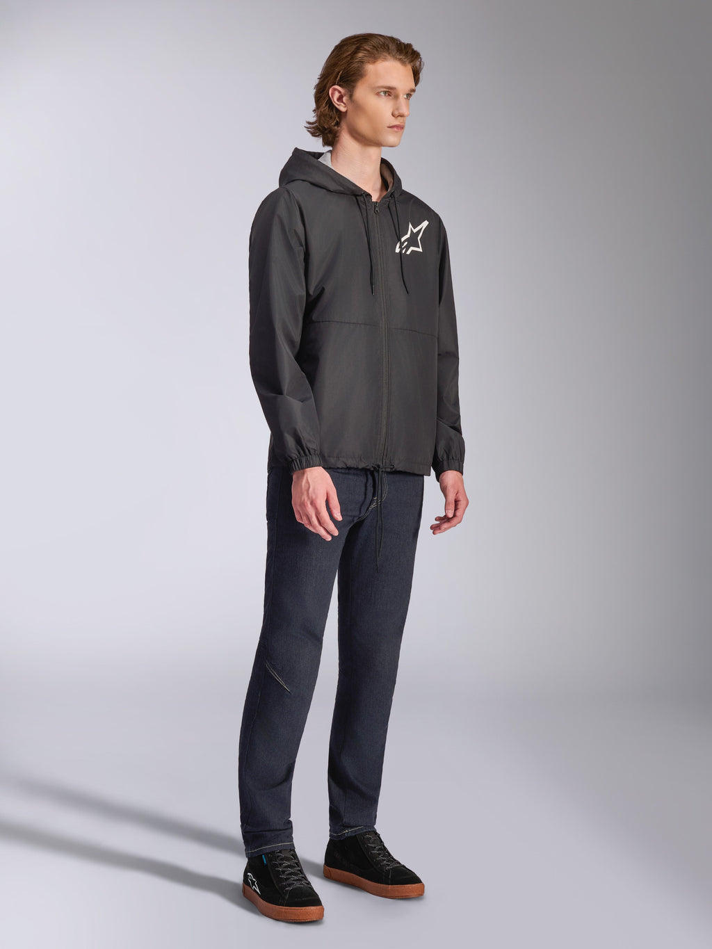 Veste coupe-vent Alpinestars Speeder Chromium, noire, coupe-vent à capuche avec fermeture éclair avant, logo étoile Alpinestars blanc sur la poitrine gauche, streetwear de sport automobile décontracté, portée avec un pantalon sombre et des baskets noires en studio.