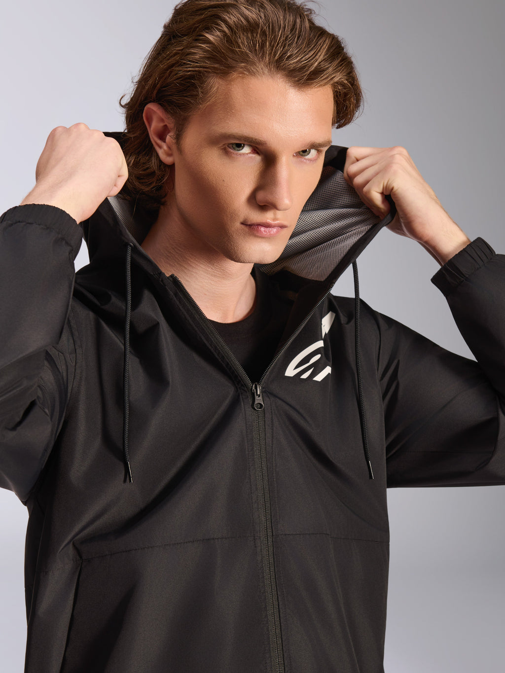 Veste coupe-vent Alpinestars Speeder Chromium, noire, portée par un mannequin homme tenant la capuche, avec un logo blanc sur la poitrine, une doublure de capuche en mesh et une fermeture éclair intégrale, streetwear décontracté inspiré des sports mécaniques