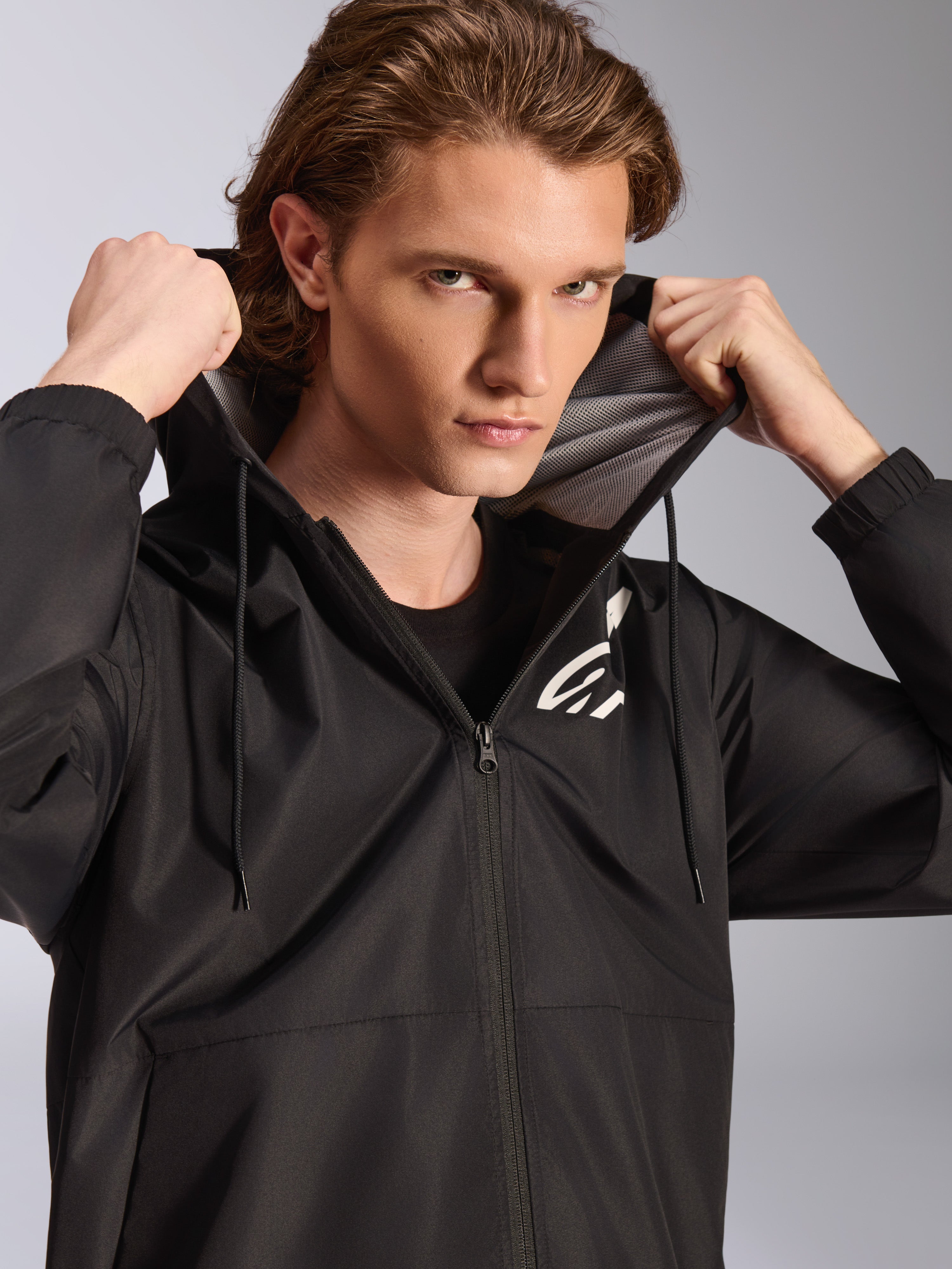 Veste coupe-vent Alpinestars Speeder Chromium, noire, portée par un mannequin homme tenant la capuche, avec un logo blanc sur la poitrine, une doublure de capuche en mesh et une fermeture éclair intégrale, streetwear décontracté inspiré des sports mécaniques