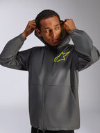 Veste coupe-vent Alpinestars Speeder Chromium, gris anthracite et jaune fluo, mannequin homme portant un coupe-vent à capuche avec cordon de serrage, logo Alpinestars jaune fluo sur la poitrine et doublure de capuche jaune, streetwear décontracté d'inspiration sport mécanique
