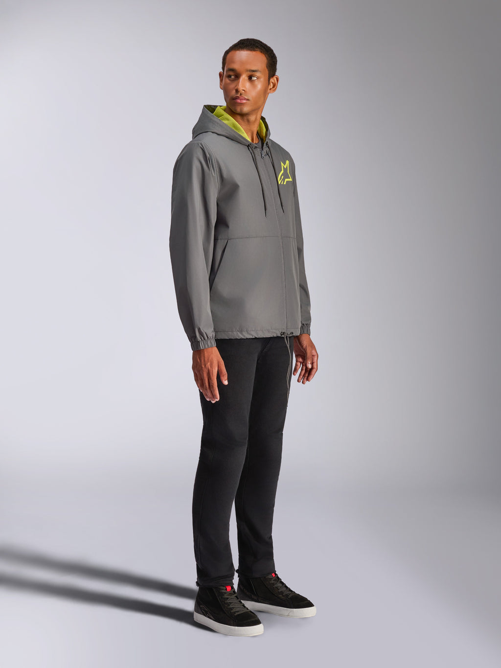 Veste coupe-vent Alpinestars Speeder Chromium, anthracite avec accents jaune fluo, mannequin homme portant un coupe-vent à capuche, logo étoile jaune sur la poitrine, associé à un pantalon noir et des baskets noires, streetwear décontracté inspiré du sport automobile