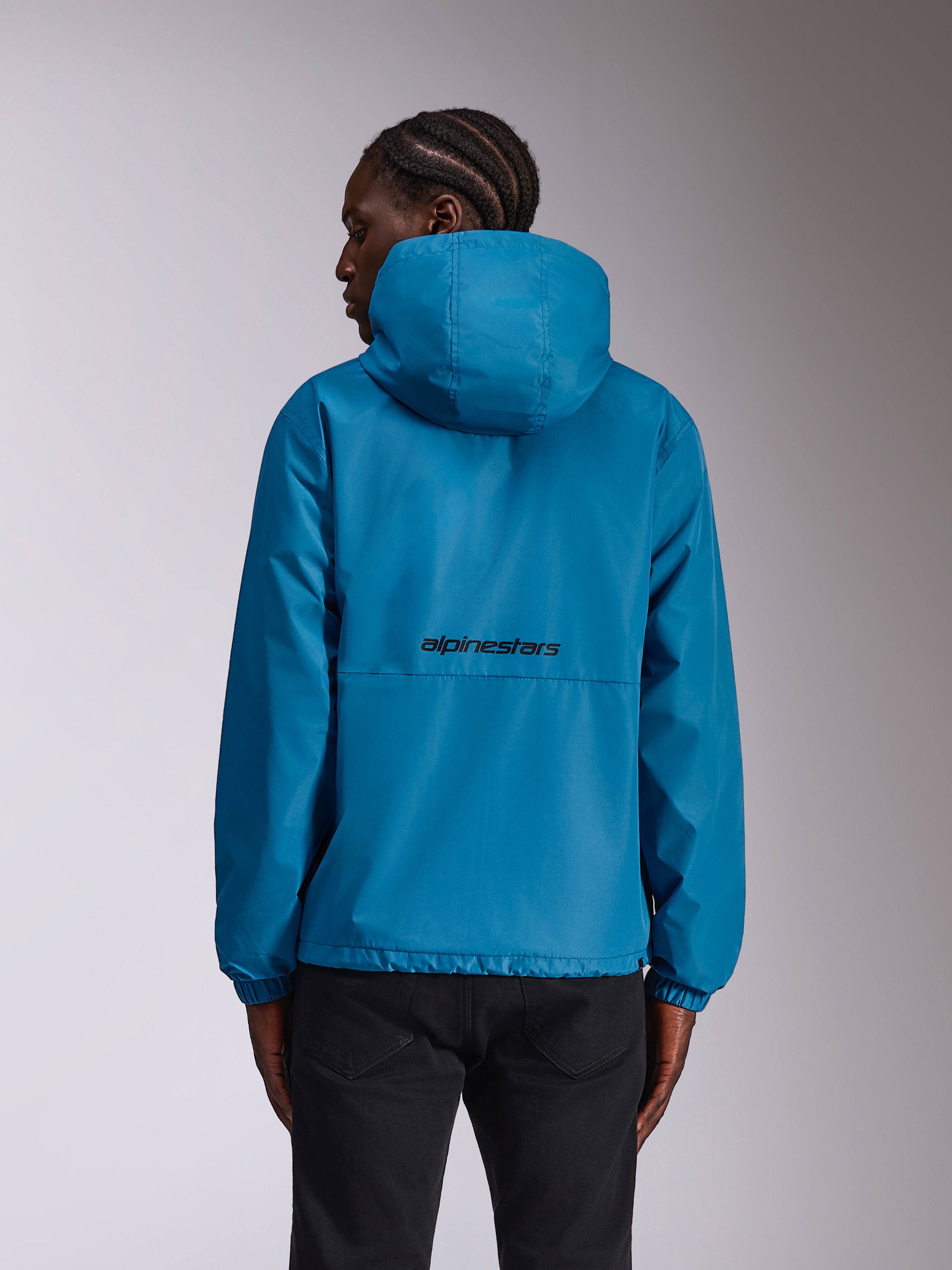 Veste coupe-vent Alpinestars Speeder Chromium, bleue, vue de dos sur mannequin montrant le logo textuel noir à l'arrière, coupe-vent léger à capuche avec poignets élastiques, vêtement de sport décontracté