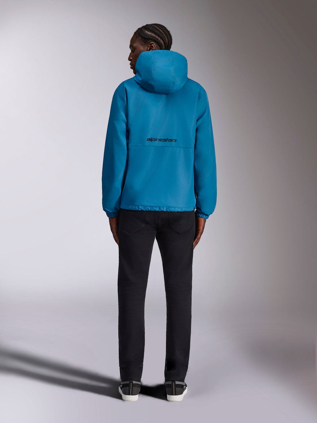 Veste coupe-vent Alpinestars Speeder Chromium, coupe-vent bleu, logo Alpinestars noir au dos, design à capuche, poignets élastiques, vêtement de sport décontracté, modèle vu de dos portant un pantalon noir