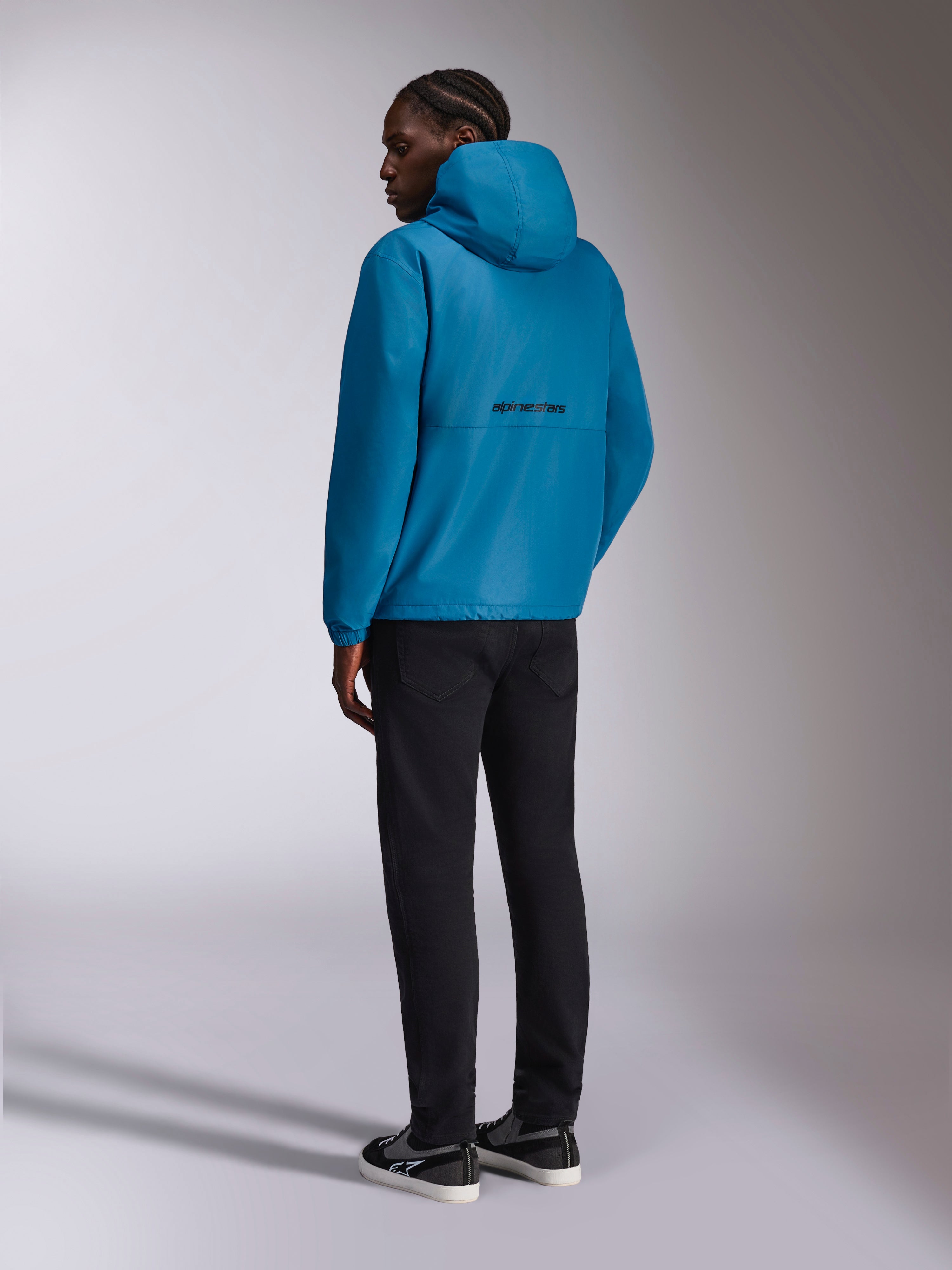 Veste coupe-vent Alpinestars Speeder Chromium, coupe-vent à capuche, bleu, vue arrière présentant le logo Alpinestars noir au dos, portée avec un pantalon noir et des baskets noires et blanches, streetwear décontracté d'inspiration sport mécanique