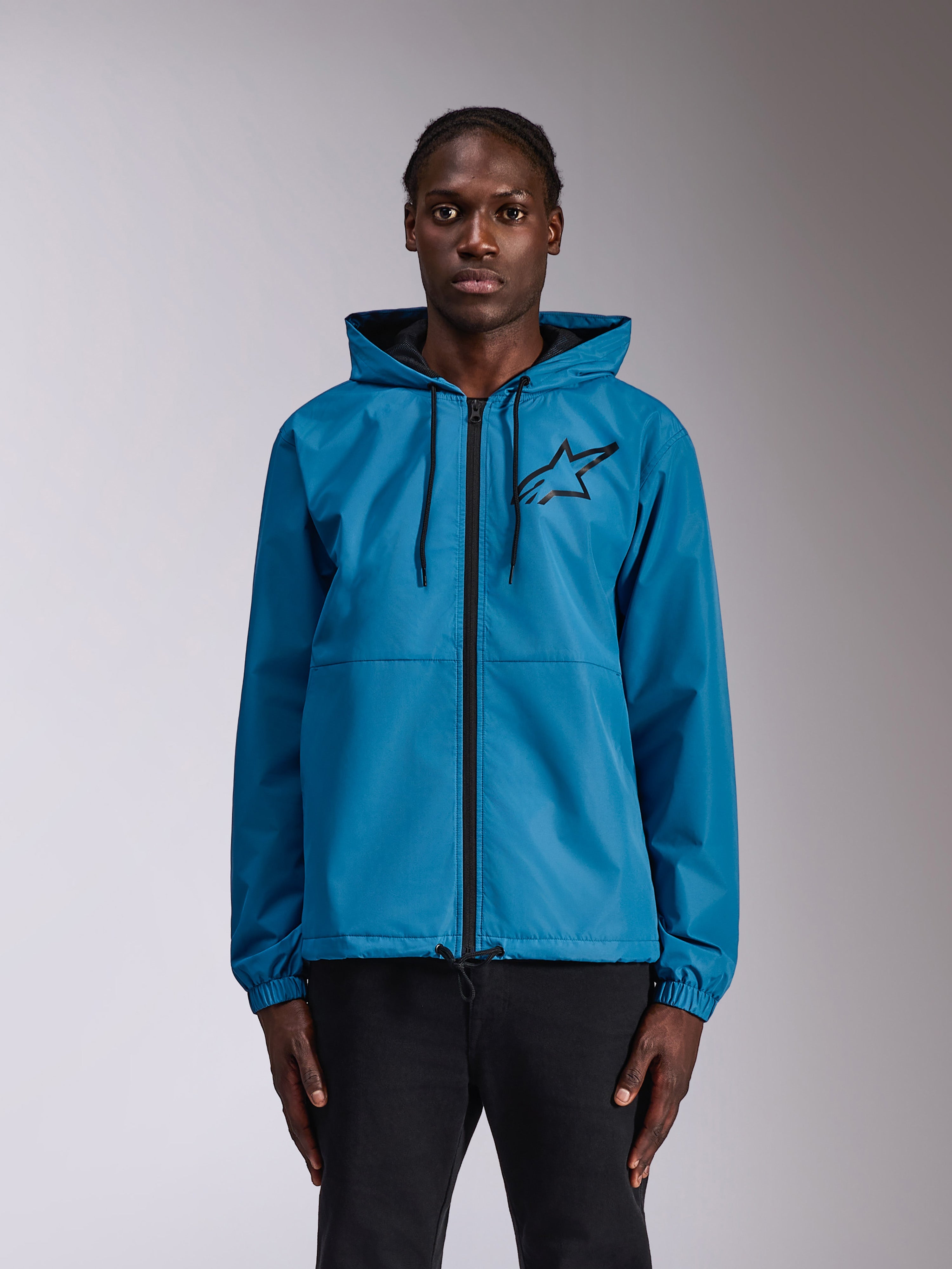 Veste coupe-vent Alpinestars Speeder Chromium, coupe-vent bleu à capuche avec logo Astars noir sur la poitrine, fermeture éclair intégrale à l'avant, poignets élastiques et capuche avec cordon de serrage, portée par un mannequin comme vêtement sportswear décontracté.
