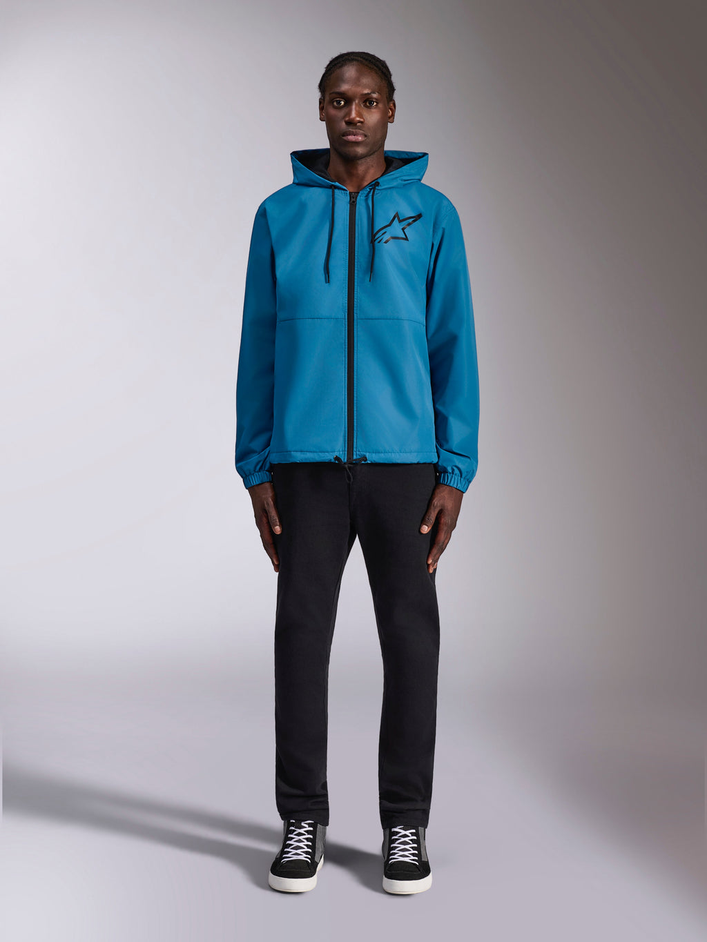 Modèle portant la veste coupe-vent Alpinestars Speeder Chromium en bleu, dotée d'une capuche avec cordons de serrage noirs, d'une fermeture éclair frontale noire et d'un logo Astars noir sur la poitrine, associée à un pantalon et des baskets noirs, style streetwear motorsport décontracté