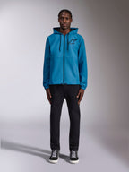 Modèle portant la veste coupe-vent Alpinestars Speeder Chromium en bleu, dotée d'une capuche avec cordons de serrage noirs, d'une fermeture éclair frontale noire et d'un logo Astars noir sur la poitrine, associée à un pantalon et des baskets noirs, style streetwear motorsport décontracté