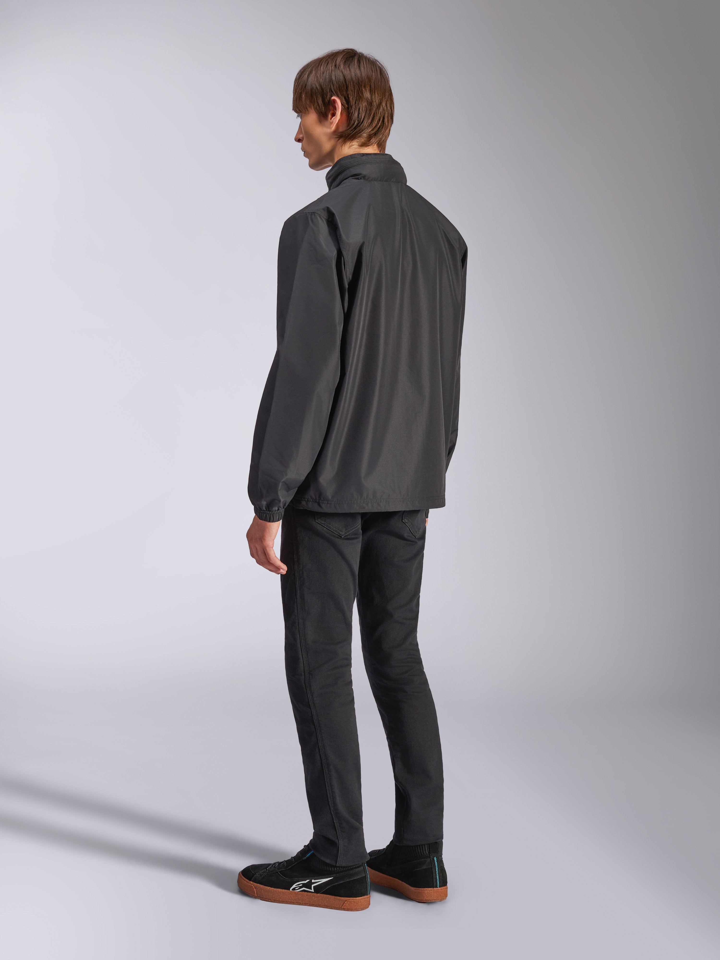 Veste Alpinestars Overhaul Racer, noire, vue de dos aux trois-quarts, portée par un mannequin avec un jean noir et des baskets noires, veste de pluie légère résistante à l'eau pour un style de vie décontracté inspiré du sport automobile
