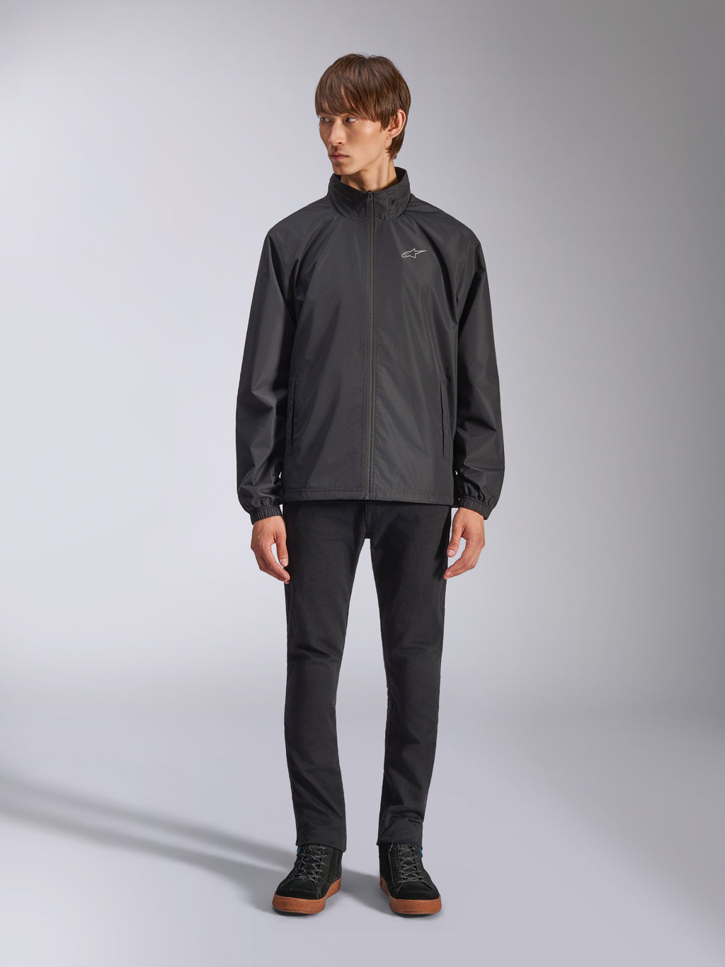 Veste Alpinestars Overhaul Racer, veste de pluie noire pour homme, vue en pied sur un mannequin avec pantalon noir et baskets décontractées, dotée d'un petit logo blanc sur la poitrine, d'un tissu résistant à l'eau et d'un design décontracté inspiré du sport automobile
