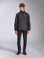 Veste Alpinestars Overhaul Racer, veste de pluie noire pour homme, vue en pied sur un mannequin avec pantalon noir et baskets décontractées, dotée d'un petit logo blanc sur la poitrine, d'un tissu résistant à l'eau et d'un design décontracté inspiré du sport automobile