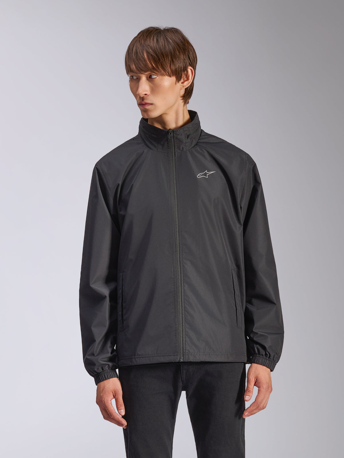 Alpinestars Overhaul Racer Jacket, veste de pluie noire pour homme, avec col montant et fermeture éclair frontale intégrale, petit logo blanc sur la poitrine, poignets élastiques, tissu lisse résistant à l'eau, design minimaliste inspiré du sport automobile