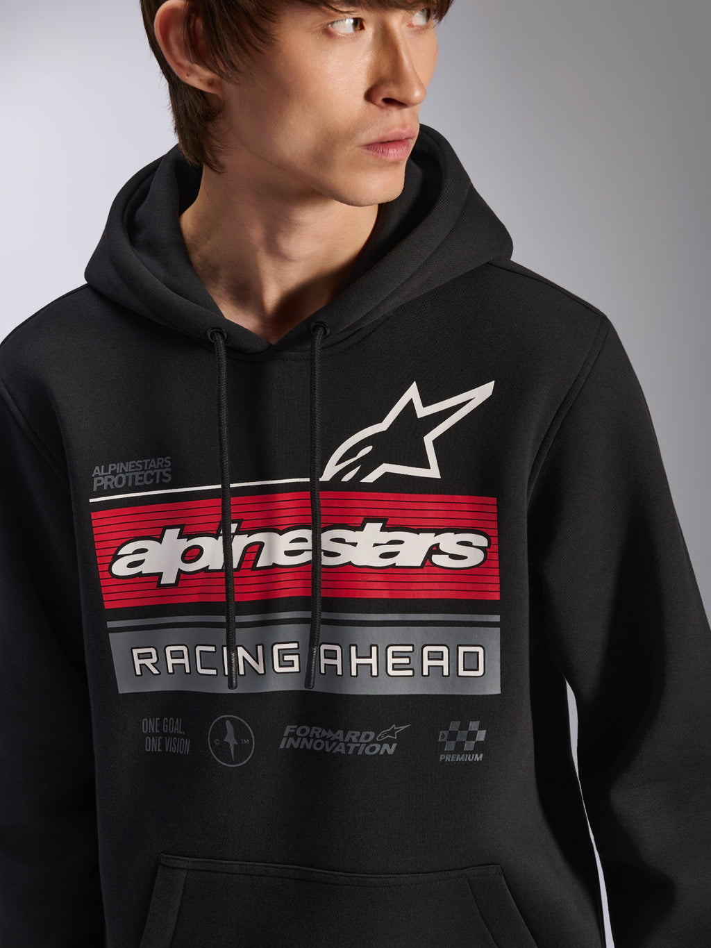 Sweat à capuche Alpinestars Harken, sweat en polaire noir porté par un mannequin, doté d'un grand graphisme sur la poitrine avec des bandes rouges et grises, logo Alpinestars blanc et logo étoile, inscription « Racing Ahead », streetwear décontracté moderne d'inspiration sport mécanique.