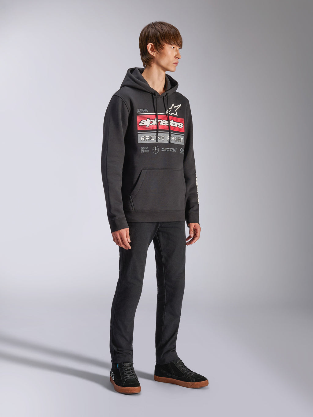 Sweat à capuche Harken Alpinestars, sweat à capuche noir en polaire, doté d'un grand graphisme sur la poitrine avec lettrage Alpinestars rouge et logo étoile, poche kangourou, porté par un mannequin avec un pantalon et des baskets noirs, style streetwear décontracté motorsport