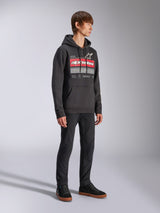 Sweat à capuche Harken Alpinestars, sweat à capuche noir en polaire, doté d'un grand graphisme sur la poitrine avec lettrage Alpinestars rouge et logo étoile, poche kangourou, porté par un mannequin avec un pantalon et des baskets noirs, style streetwear décontracté motorsport