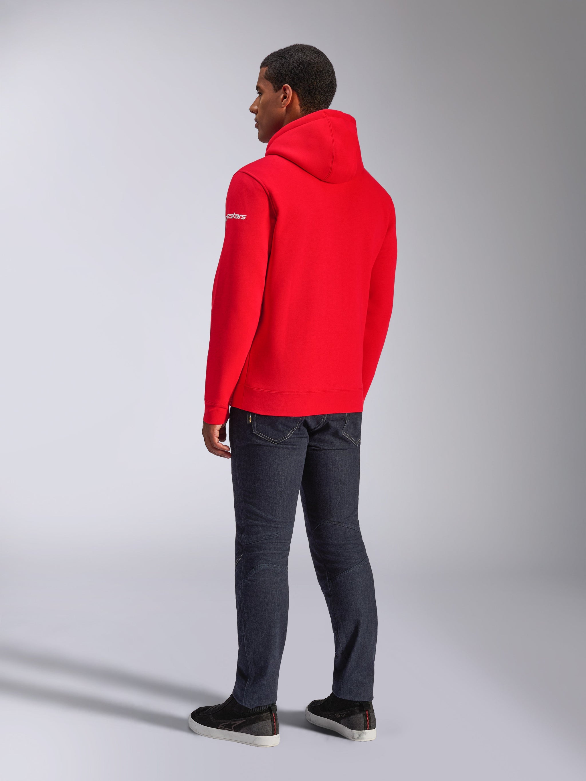 Sweat à capuche Frontal Alpinestars, sweat à capuche en polaire rouge, vue de dos montrant le logo wordmark blanc sur la manche, porté avec un pantalon en jean foncé et des chaussures noires, style streetwear décontracté motorsport