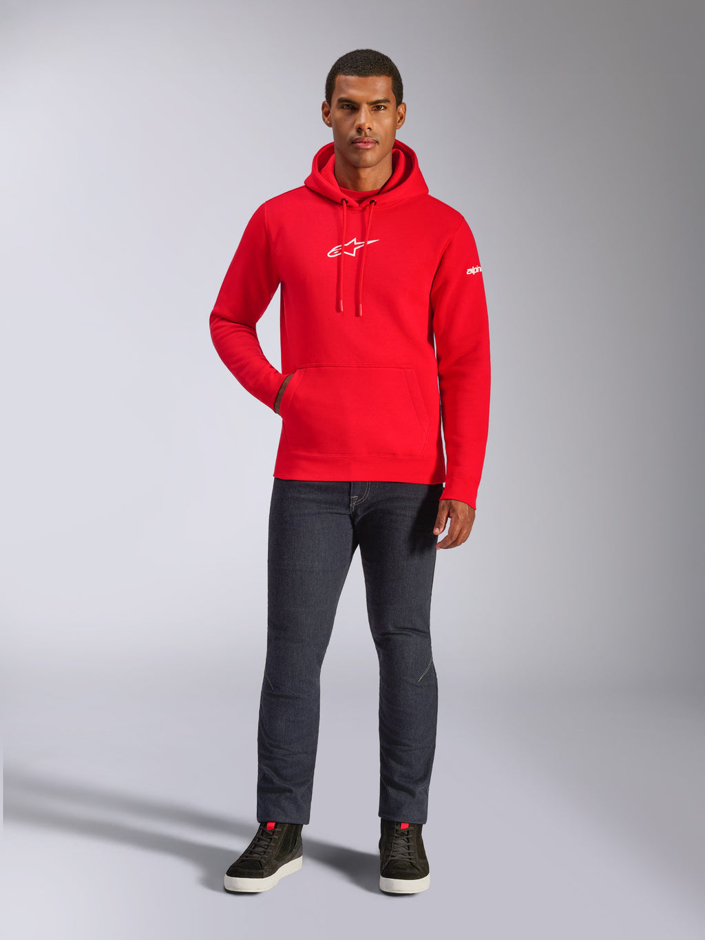 Modèle masculin portant le sweat à capuche Frontal d'Alpinestars en rouge, sweat à capuche en polaire à enfiler avec logo Astars blanc sur la poitrine, poche kangourou, capuche à cordon de serrage, associé à un jean gris foncé et des baskets montantes noires, style streetwear motorsport décontracté