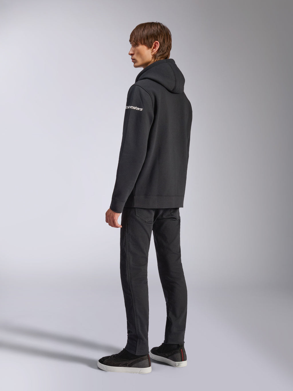 Sweat à capuche Alpinestars Frontal, streetwear décontracté, noir, vue de dos latérale montrant le logo wordmark blanc sur la manche, porté avec un pantalon noir et des baskets, design moderne inspiré du sport automobile