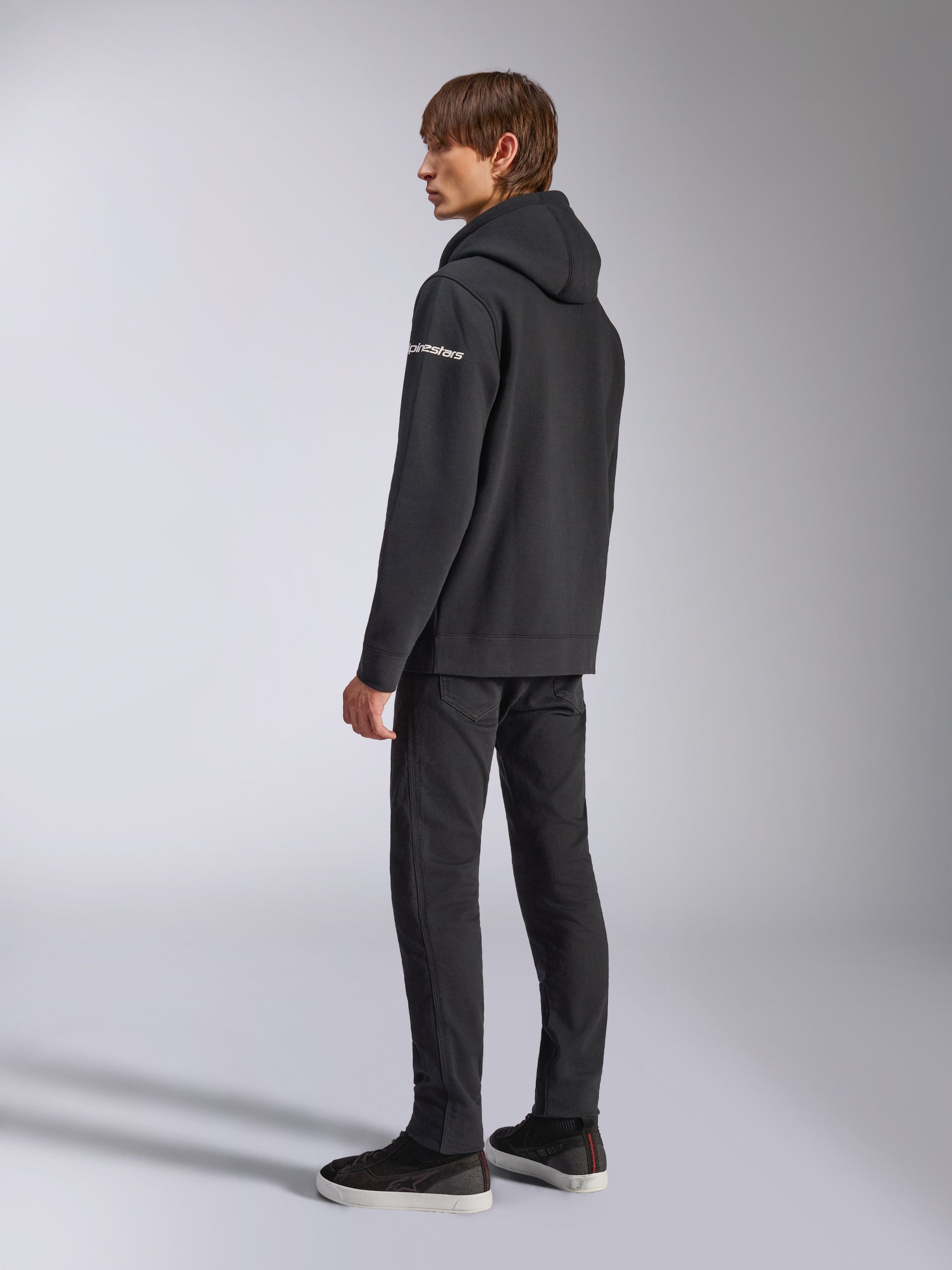 Sweat à capuche Alpinestars Frontal, streetwear décontracté, noir, vue de dos latérale montrant le logo wordmark blanc sur la manche, porté avec un pantalon noir et des baskets, design moderne inspiré du sport automobile