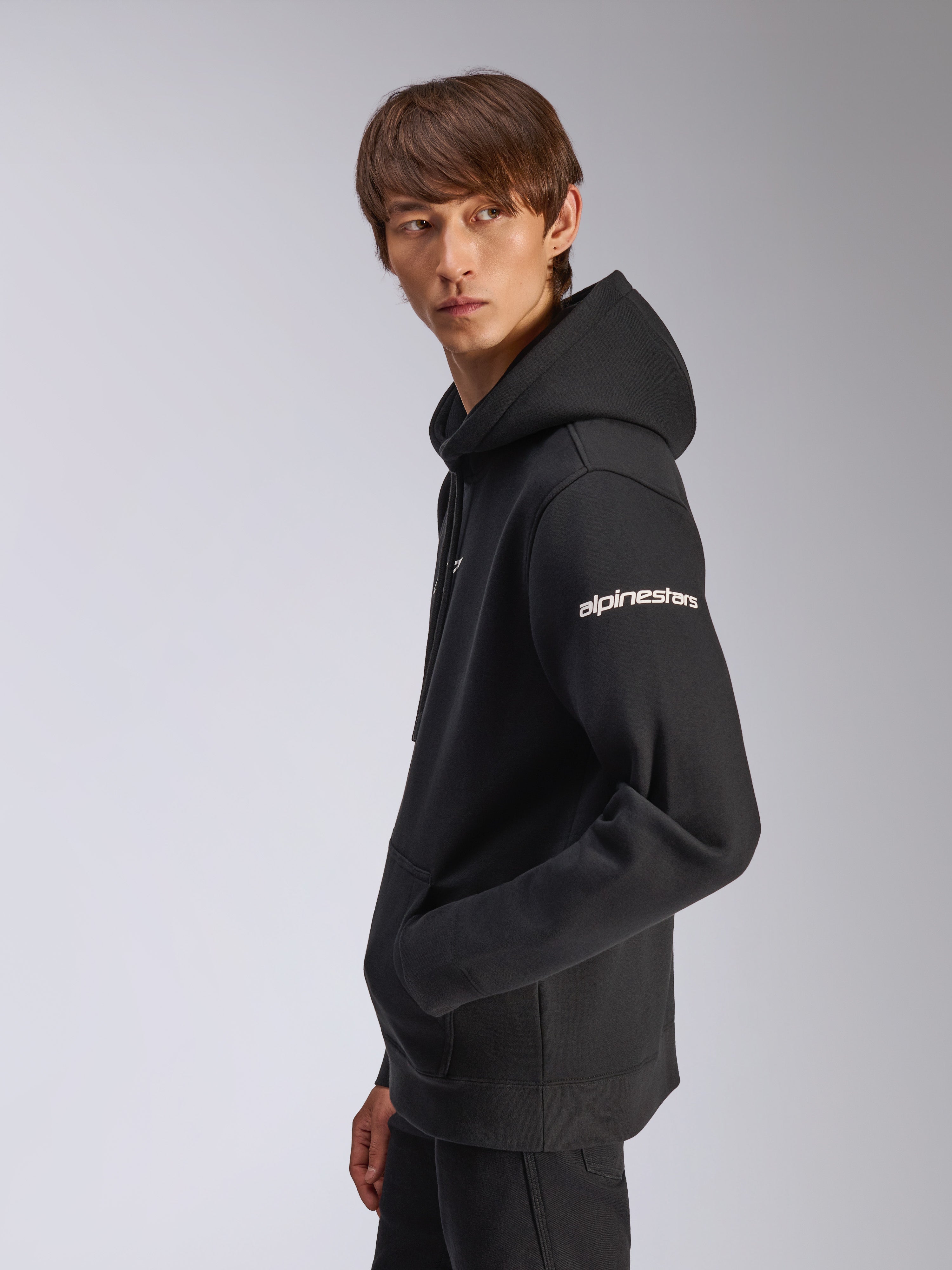 Sweat à capuche Frontal Alpinestars, polaire noire à enfiler, mannequin de profil, logo Alpinestars blanc sur la manche, poche kangourou, streetwear décontracté d'inspiration sport automobile