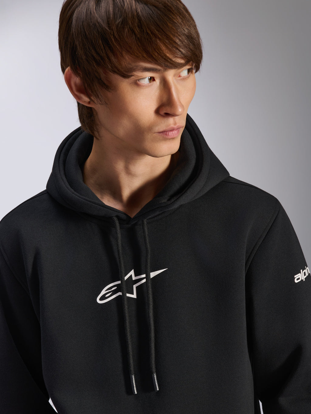 Sweat à capuche Frontal Alpinestars, sweat à capuche noir en polaire à enfiler, logo Astar blanc sur la poitrine et logo sur la manche, capuche à cordon, porté par un mannequin homme, style streetwear décontracté motorsport