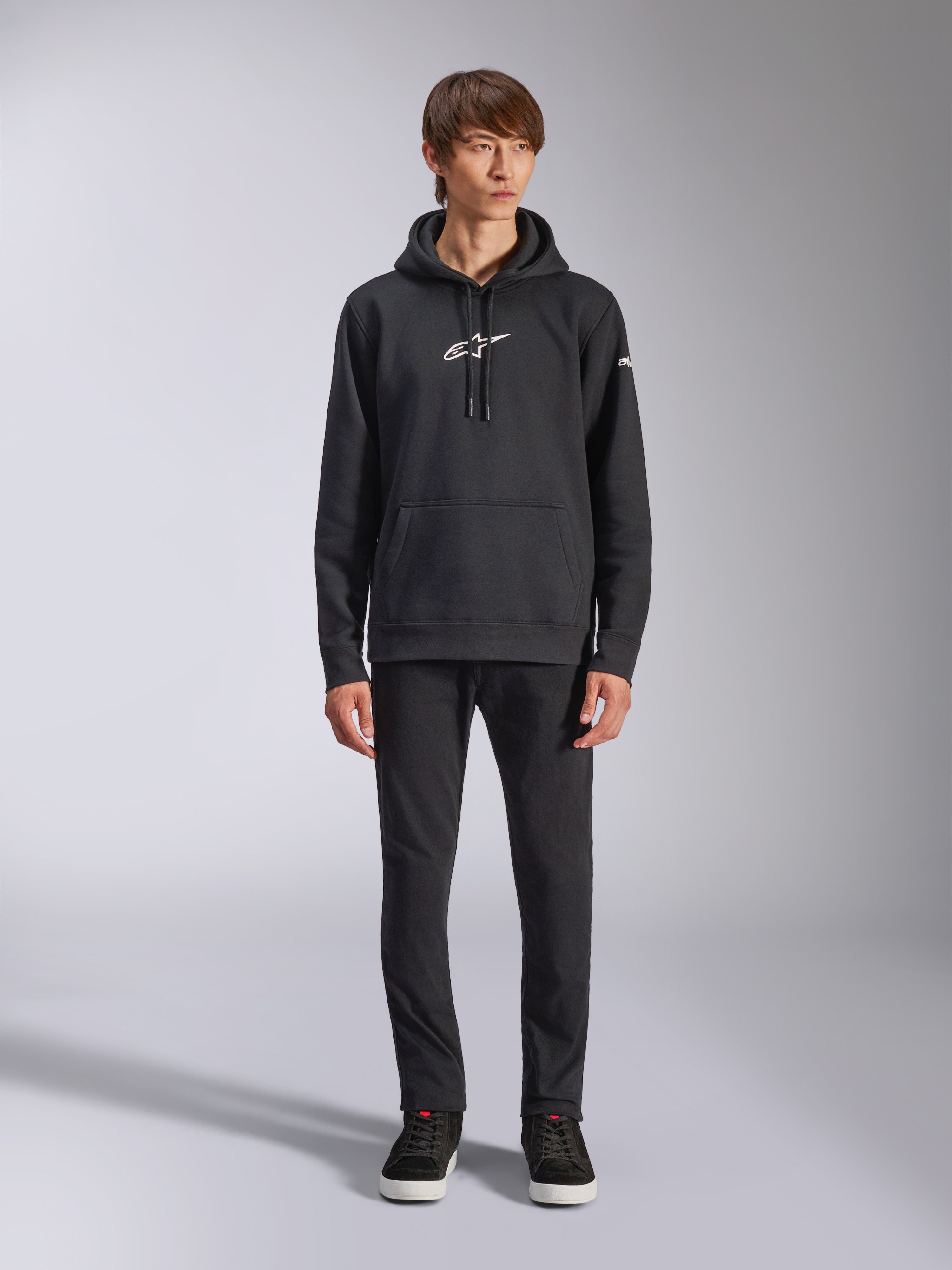 Sweat à capuche Alpinestars Frontal, sweat à capuche noir en polaire, logo Alpinestars blanc sur la poitrine et la manche, poche kangourou, capuche à cordon de serrage, porté avec un pantalon noir, design décontracté inspiré des sports mécaniques