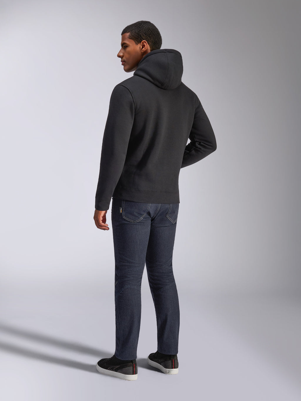 Alpinestars Blaze V3 Hoodie, vue de dos d'un homme portant le sweat à capuche noir, associé à un jean foncé et des baskets noires, style streetwear décontracté inspiré du sport automobile