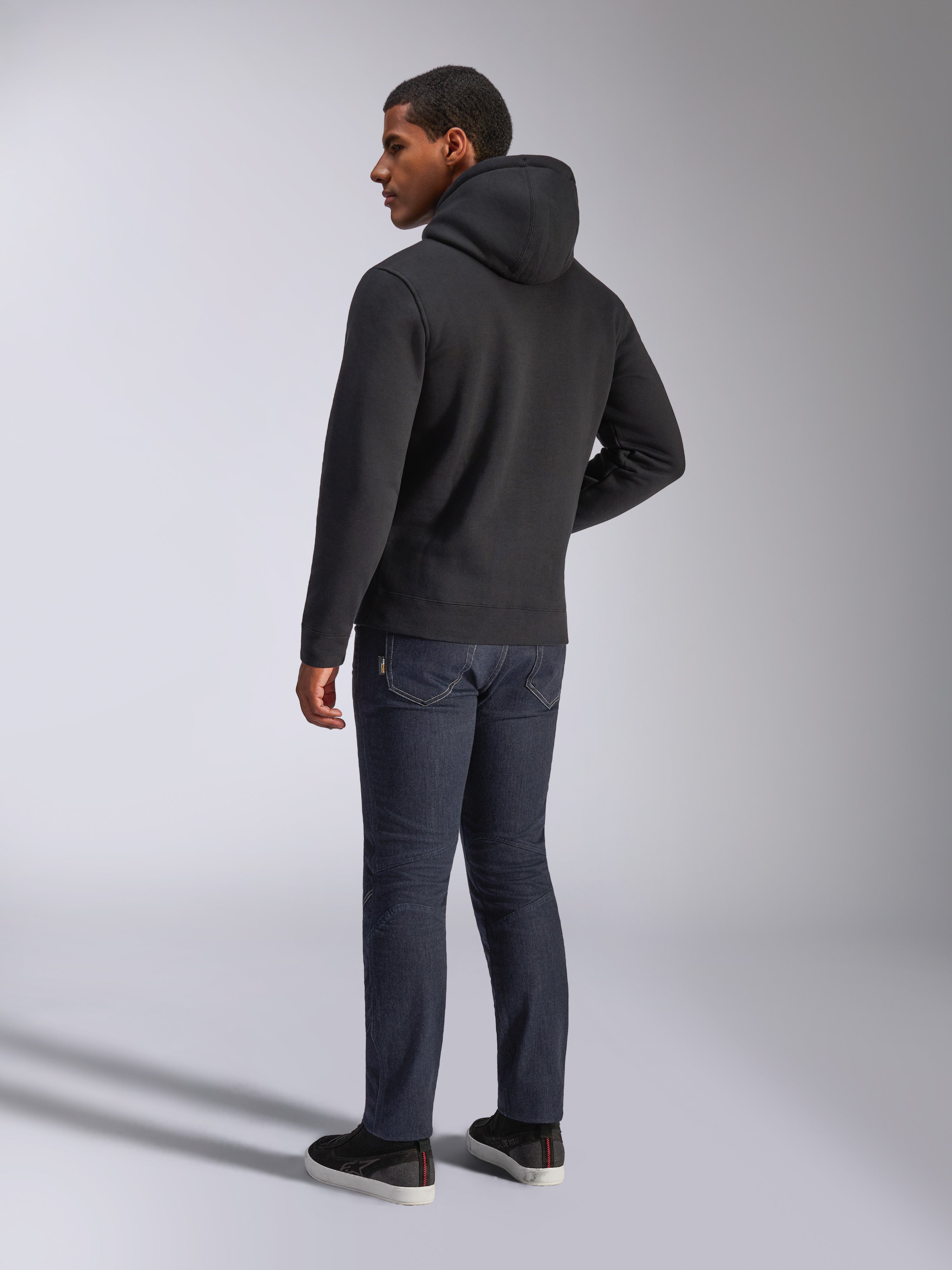 Alpinestars Blaze V3 Hoodie, vue de dos d'un homme portant le sweat à capuche noir, associé à un jean foncé et des baskets noires, style streetwear décontracté inspiré du sport automobile