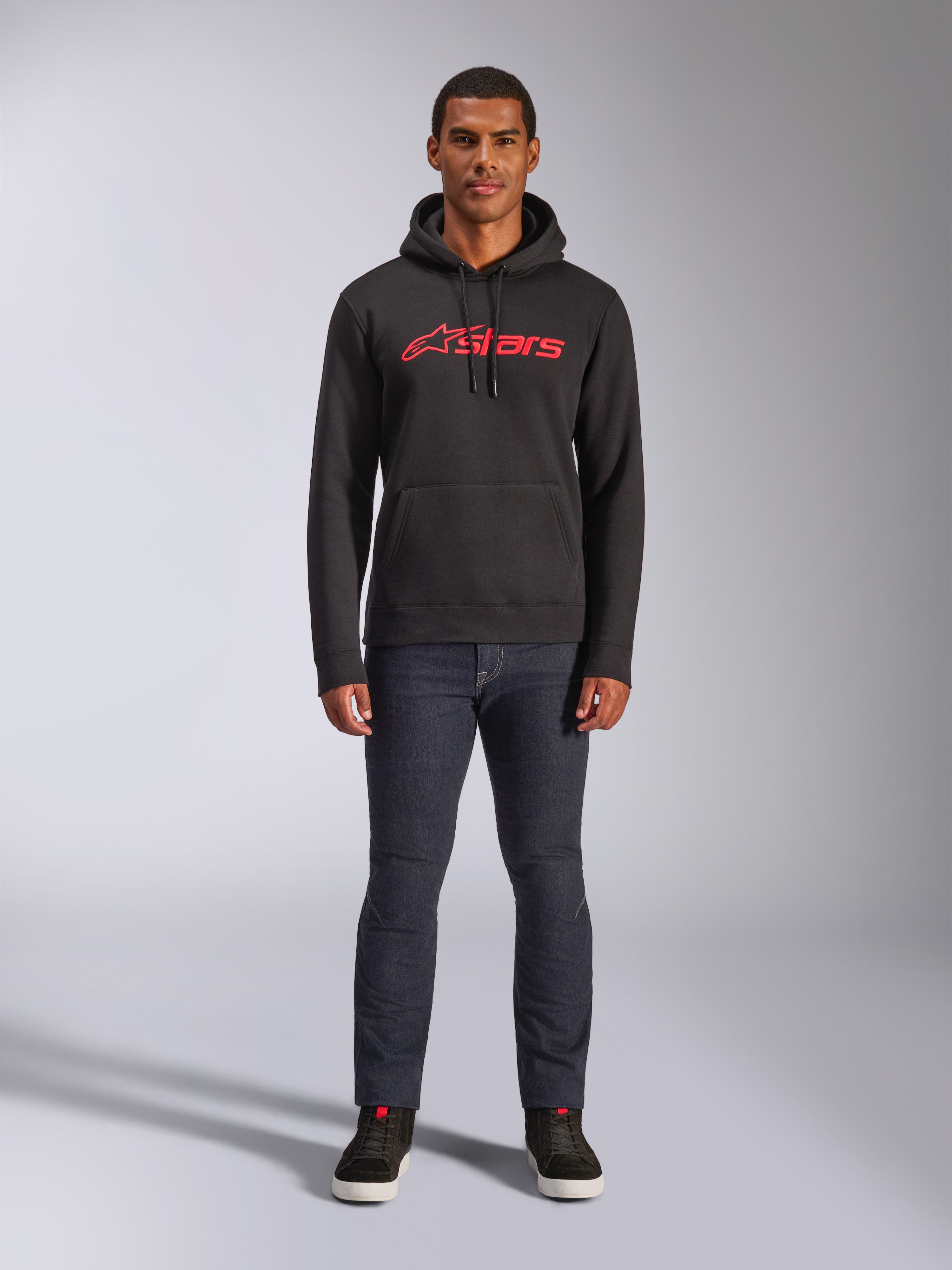 Sweat à capuche Alpinestars Blaze V3, style streetwear décontracté, noir avec logo Astars rouge, vue de face, porté par un mannequin avec un pantalon en denim foncé et des baskets noires, design moderne en polaire inspiré des sports mécaniques.