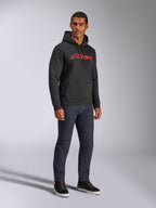 Sweat à capuche Alpinestars Blaze V3, polaire noire avec logo Alpinestars rouge sur la poitrine, porté par un mannequin homme avec un jean en denim foncé et des baskets noires, streetwear décontracté inspiré des sports mécaniques