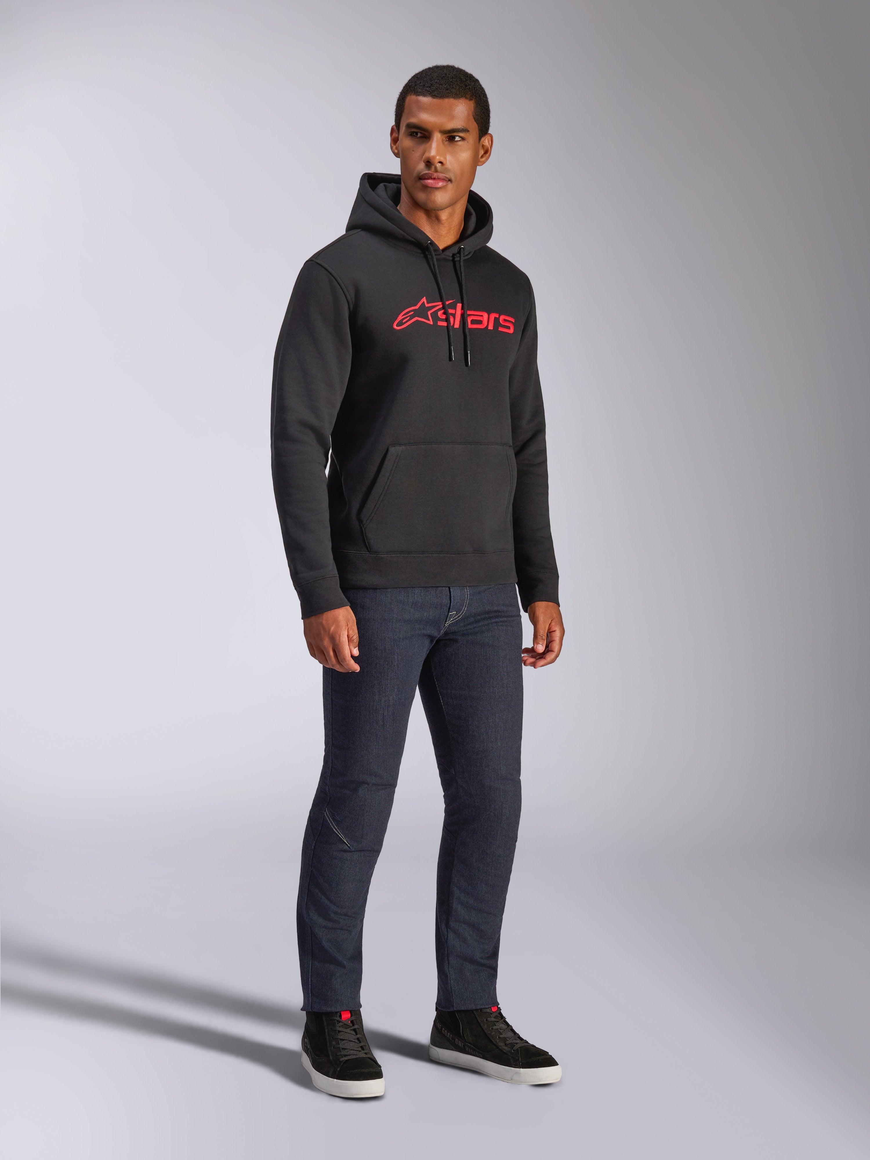 Sweat à capuche Alpinestars Blaze V3, polaire noire avec logo Alpinestars rouge sur la poitrine, porté par un mannequin homme avec un jean en denim foncé et des baskets noires, streetwear décontracté inspiré des sports mécaniques