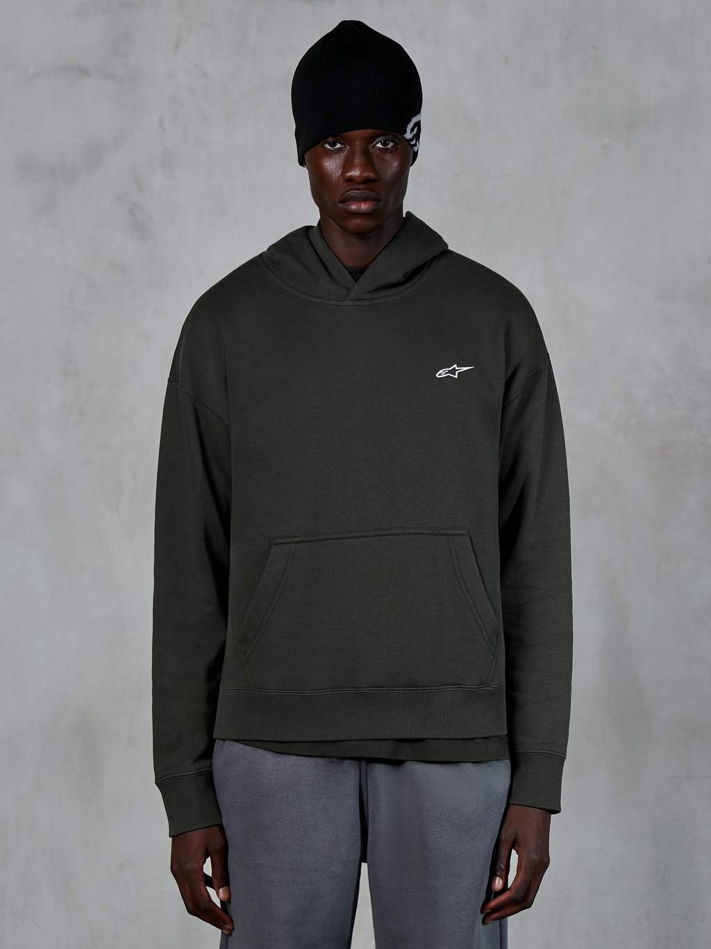 Sweat à capuche Alpinestars Essentials Ageless, style streetwear décontracté, pullover Off Black avec un petit logo Alpinestars blanc sur la poitrine, porté avec un bonnet noir, design minimaliste avec une poche kangourou à l'avant et finition délavée