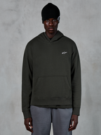 Sweat à capuche Alpinestars Essentials Ageless, style streetwear décontracté, pullover Off Black avec un petit logo Alpinestars blanc sur la poitrine, porté avec un bonnet noir, design minimaliste avec une poche kangourou à l'avant et finition délavée