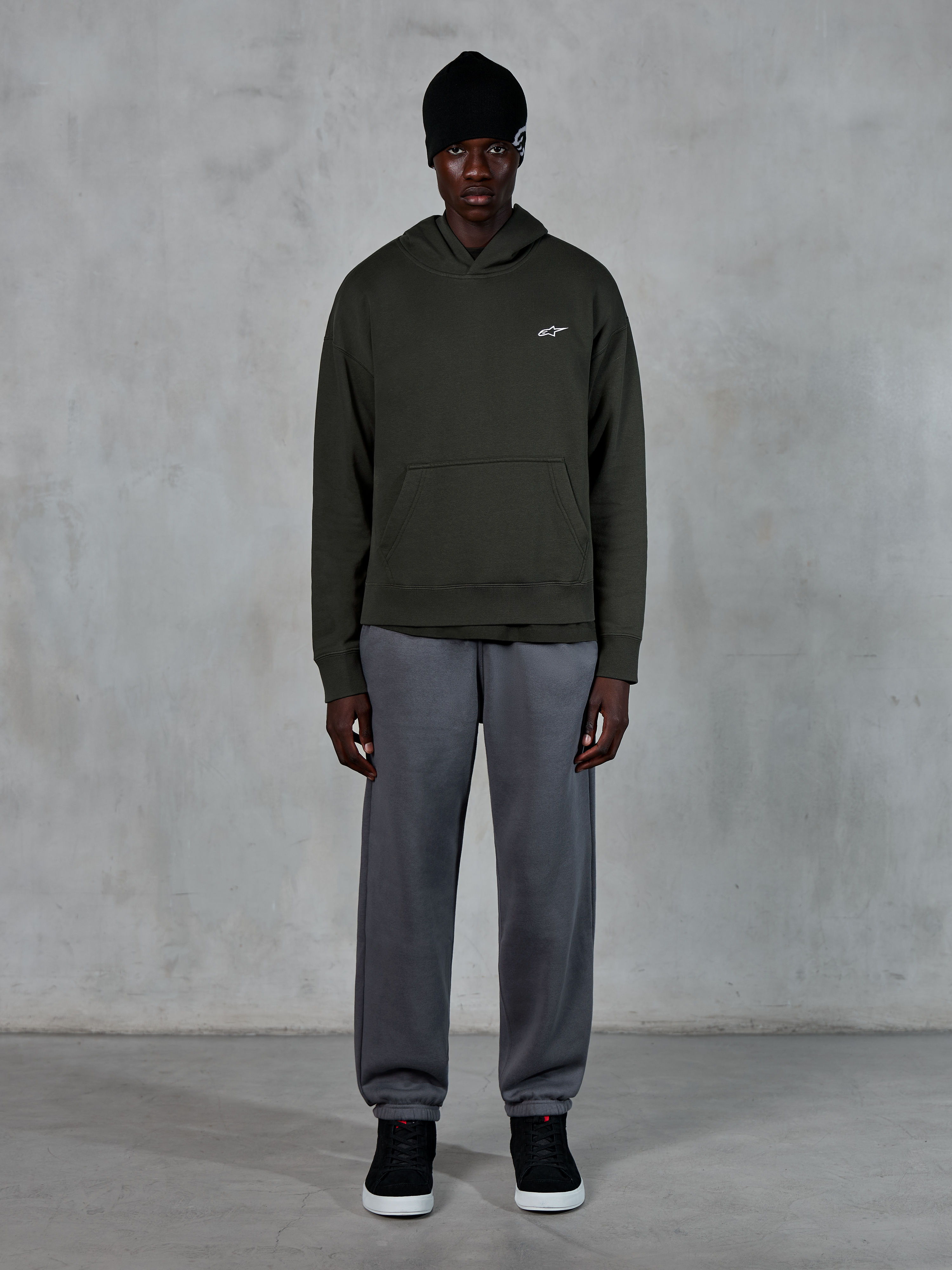 Modèle portant le sweat à capuche Alpinestars Essentials Ageless, streetwear décontracté, Off Black avec un petit logo blanc sur la poitrine, associé à un jogging gris, un bonnet noir et des baskets noires, design minimaliste inspiré du sport automobile