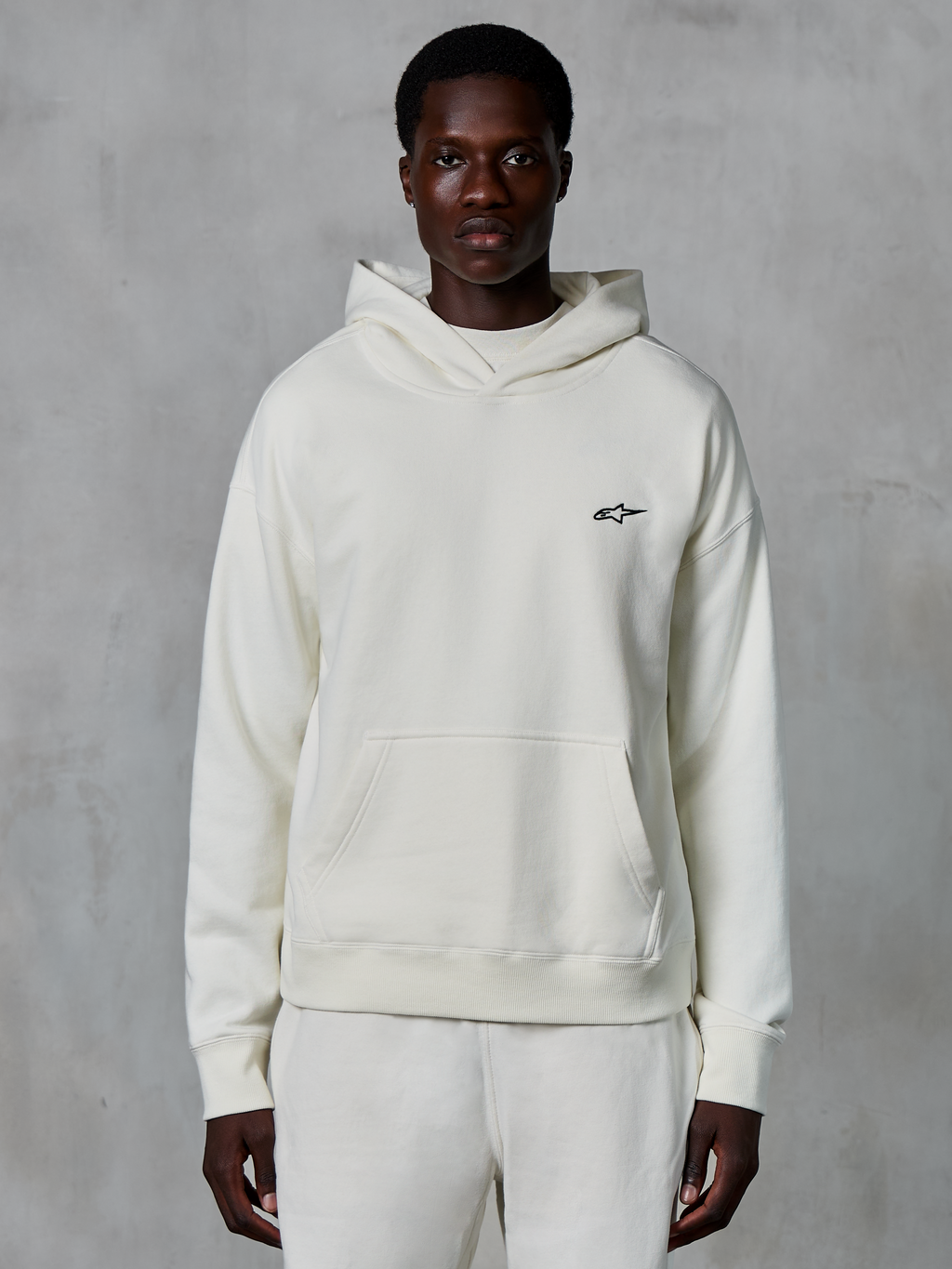 Sweat à capuche Alpinestars Essentials Ageless, blanc cassé, porté par un mannequin homme, petit logo noir sur la poitrine, poche kangourou frontale, manches longues, streetwear décontracté inspiré du sport automobile