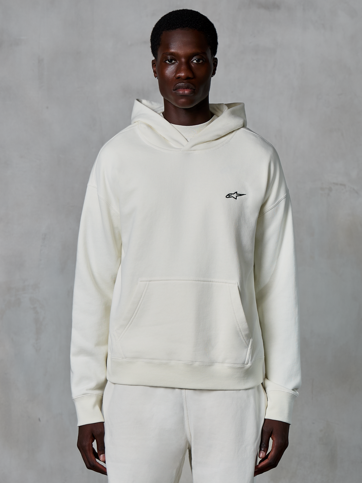 Sweat à capuche Alpinestars Essentials Ageless, blanc cassé, porté par un mannequin homme, petit logo noir sur la poitrine, poche kangourou frontale, manches longues, streetwear décontracté inspiré du sport automobile