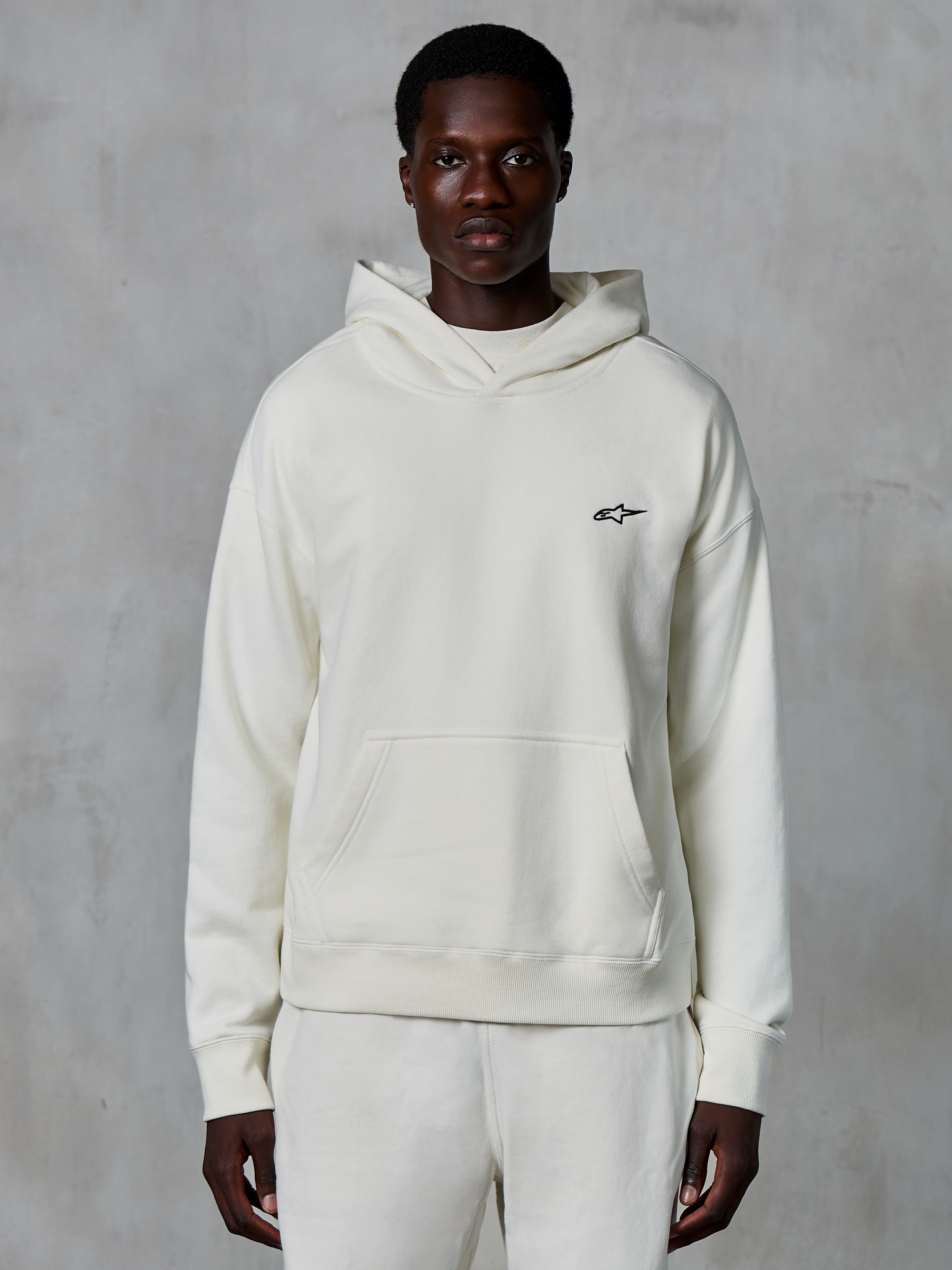 Sweat à capuche Alpinestars Essentials Ageless, blanc cassé, porté par un mannequin homme, petit logo noir sur la poitrine, poche kangourou frontale, manches longues, streetwear décontracté inspiré du sport automobile