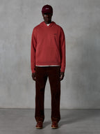 Sweat à capuche Essentials Ageless Alpinestars, sweat à capuche sportswear décontracté rouge brique avec finition délavée à la pierre, arborant un petit logo Alpinestars noir sur la poitrine et une poche kangourou à l'avant, porté par un mannequin avec une casquette bordeaux et un pantalon en velours côtelé marron pour un look streetwear épuré.