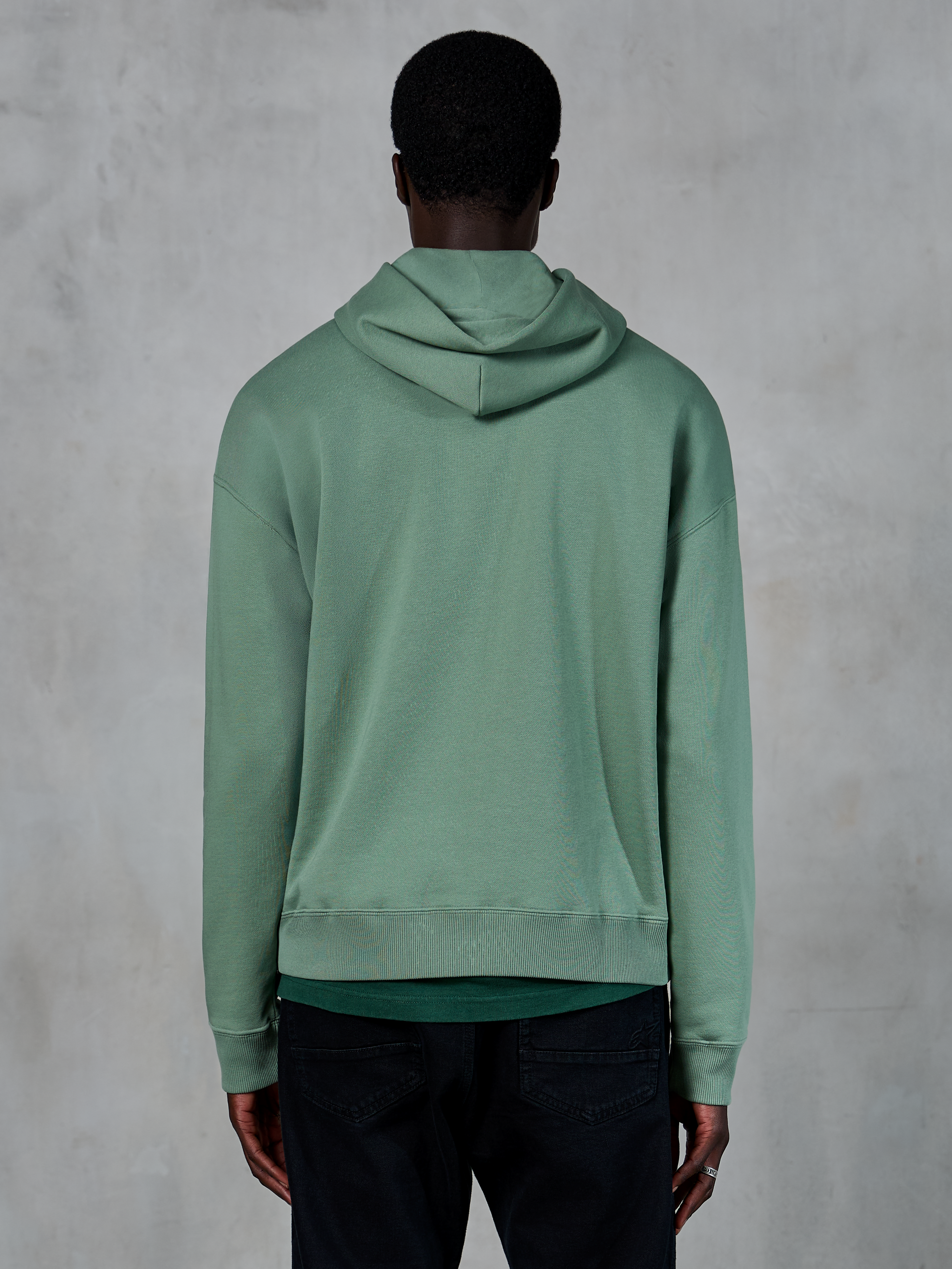 Sweat à capuche Alpinestars Essentials Ageless, sportswear décontracté, vert Cactus, vue de dos sur modèle, finition lavage à la pierre, porté avec un pantalon noir, design lifestyle minimaliste